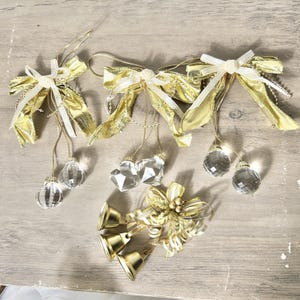 May include: Gold and white decorative bows with crystal-like ornaments and gold bells. The bows have a satin finish and are accented with white ribbon and hanging crystal pendants. The bells are small and gold.