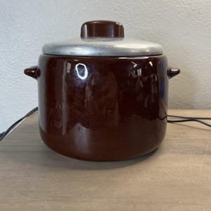 May include: A brown ceramic pot with a silver lid and a brown knob. The pot has two small handles on either side. The pot is on a wooden surface.