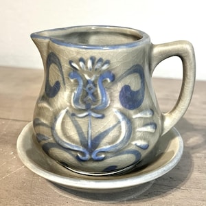 May include: A small, ceramic creamer and saucer set. The creamer is beige with blue floral designs and a handle. The saucer is a matching beige with a blue rim. The set is placed on a wooden surface.