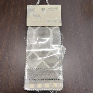 Puede incluir: Muestras de tela en una funda de plástico transparente. Presentan varios patrones grises y blancos, incluyendo diseños geométricos y texturizados. La etiqueta superior muestra el logotipo "Kasmir concepts" y el texto "Book Soft Volume 2 Tag-A-Longs Multi-Purpose Coordinates."