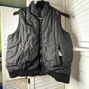 May include: Black quilted vest with a high collar and a full-length zipper. The vest has two front pockets and a ribbed hem. The vest is hanging on a black hanger.