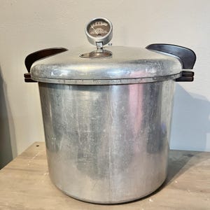 Könnte beinhalten: Ein alter Aluminium-Schnellkochtopf mit einem Messgerät oben und dunkelbraunen Griffen. Der Kocher ist zylindrisch mit einem Deckel und steht auf einer hellen Holzoberfläche.