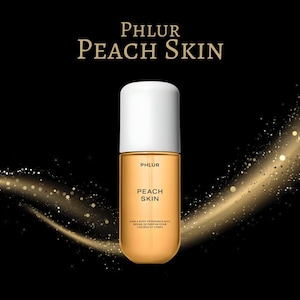 May include: A peach-colored fragrance mist bottle with a white cap, labeled "PHLUR PEACH SKIN." The bottle is set against a black background with gold glitter accents. The text "HAIR & BODY FRAGRANCE MIST BRUME DE PARFUM POUR CHEVEUX ET CORPS" is also visible.