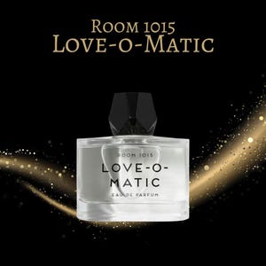 May include: A rectangular perfume bottle with a black geometric cap. The bottle is clear, filled with a light-colored liquid, and features the text "ROOM 1015 LOVE-O-MATIC EAU DE PARFUM" in black. The background is black with gold glitter.