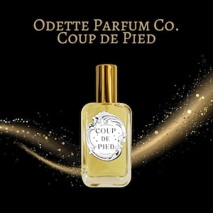 May include: A perfume bottle with a gold cap and a white label with the text "COUP DE PIED" is centered on a black background. The brand name "ODETTE PARFUM CO." is above the bottle. Gold sparkles emanate from the right side.