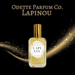 May include: A perfume bottle with a gold cap and a white label featuring the text "LAPINOU" and "ODETTE PARFUM CO." on a black background with gold glitter accents. The bottle contains a light yellow liquid.