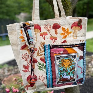 May include: Cream-colored tote bag with a whimsical mushroom and leaf print. Features a pocket with a tarot card design and decorative trim. Includes long handles and a small leather accent.