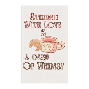 以下が含まれることがあります： ピンクと茶色で「Stirred With Love & A Dash Of Whimsy」の文字が入った白い長方形のキッチンタオル。デザインには、漫画のクロワッサンと星柄のピンクのマグカップが含まれています。