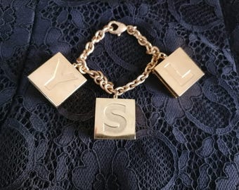 Puede incluir: Pulsera dorada con tres colgantes cuadrados, cada uno con una letra: Y, S y L. La pulsera tiene un diseño de cadena y un cierre. El fondo es una tela azul oscuro texturizada.