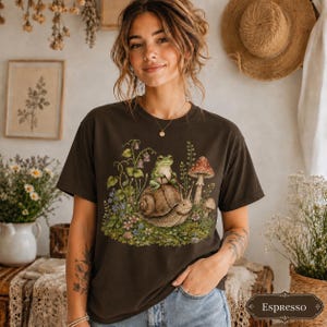Woman wearing a Comfort Colors cottagecore frog shirt featuring a cute frog riding a snail with mushrooms, wildflowers, greenery, and whimsical woodland nature details.