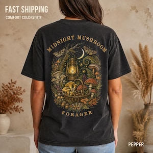 May include: A charcoal gray t-shirt featuring a back graphic of a wicker basket filled with various mushrooms, a lit lantern, and a crescent moon. The text MIDNIGHT MUSHROOM FORAGER is printed in a curved gold font around the central illustration.