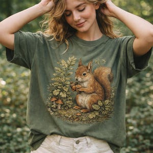 May include: An olive green t-shirt with a detailed illustration of a squirrel holding an acorn, surrounded by foliage and mushrooms. The squirrel is in shades of brown and tan, with green and gold leaves. The t-shirt has a relaxed fit.