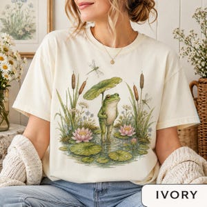 Woman wearing a Comfort Colors cottagecore frog shirt featuring a cute pond frog holding a lily pad umbrella with water lilies, dragonflies, and woodland nature details.