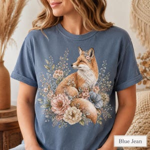 Peut inclure: T-shirt bleu jean avec une illustration détaillée d'un renard entouré de fleurs. Le renard a une fourrure orange et blanche et est orné d'une couronne florale. Les fleurs sont dans les tons de rose, bleu et crème.