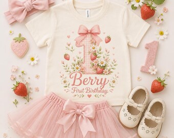 May include: A cream-colored baby outfit set featuring a t-shirt, tutu skirt, headband, and shoes. The t-shirt has a large number one with strawberries and flowers, and the text "Berry First Birthday". The set includes a pink bow and a strawberry-shaped cookie.
