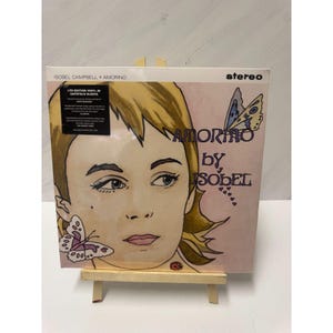 May include: A vinyl record album titled "Amorino by Isobel" with a pink cover featuring a watercolor illustration of a woman's face and butterflies. The album is displayed on a small wooden easel.