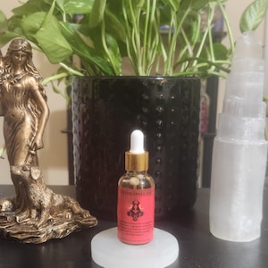 Pode incluir: Uma pequena garrafa de vidro de Lilith Spell Oil com tampa dourada e conta-gotas branco está sobre um suporte circular branco. O rótulo é vermelho com texto preto e um gráfico. Uma estátua dourada e uma torre de selenita estão em ambos os lados.