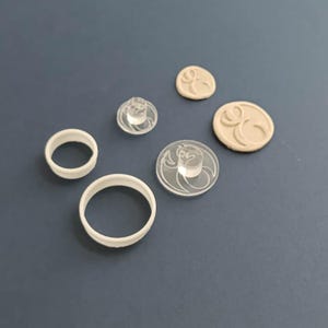 May include: A set of clear plastic cookie cutters and stamps featuring a stylized design. The set includes two round cutters, a stamp with a handle, and two impressions of the stamp in clay.