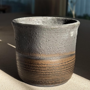Könnte beinhalten: Eine dunkelgraue Keramiktasse mit einem braunen Band um die Mitte. Die Tasse hat eine raue, strukturierte Oberfläche.