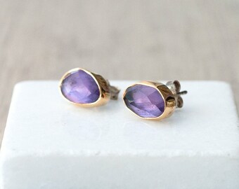 Peut inclure: Une paire de boucles d'oreilles à clous avec des pierres précieuses violettes irrégulières et facettées, serties dans des montures dorées. Les boucles d'oreilles sont présentées sur une surface blanche et rectangulaire.