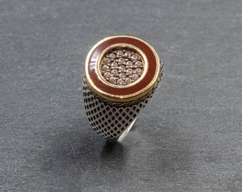 May include: A silver ring with a round, red-enameled top featuring a cluster of small, clear stones.
