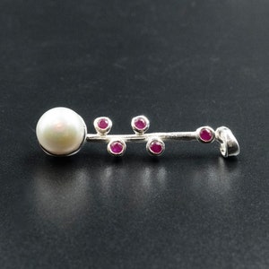 May include: A silver pendant featuring a large, round, white pearl at one end and a series of small, round, red gemstones along a linear design. The pendant has a loop for a chain.