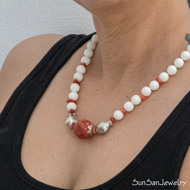 May include: A white and red beaded necklace with a large red gemstone pendant and silver accents. The necklace is worn by a person with a black tank top.