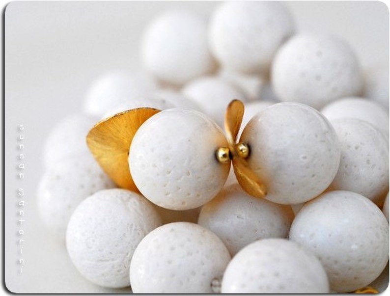 Puede incluir: Un collar de cuentas de coral blanco con detalles dorados. Las cuentas son redondas y tienen una superficie texturizada. El collar est&aacute; enhebrado en un cord&oacute;n blanco.