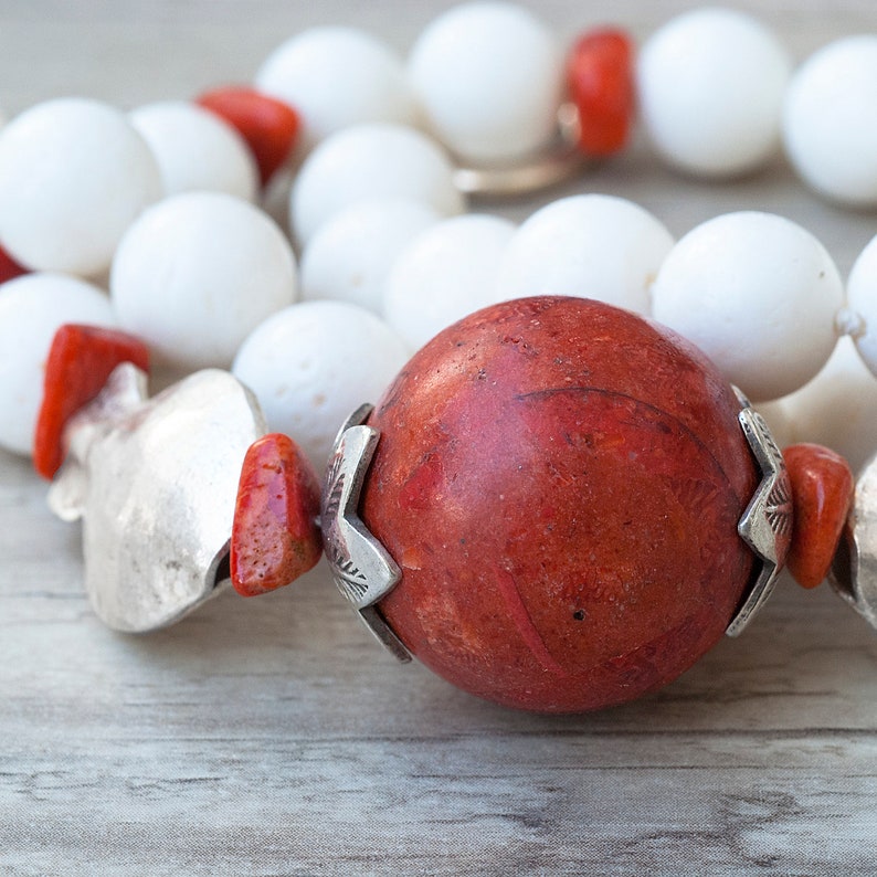 May include: A close-up of a red coral bead necklace with silver accents. The necklace is made of white and red beads and has a large, round red coral bead in the center.