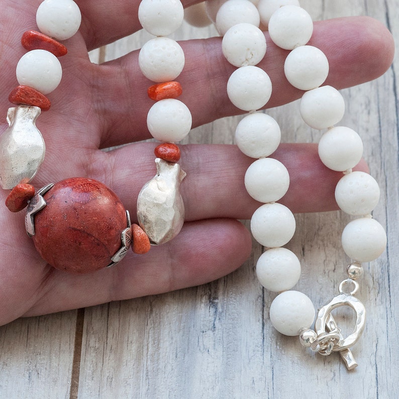 May include: A white coral bead necklace with silver pomegranate charms and red coral accents. The necklace is held in a hand.
