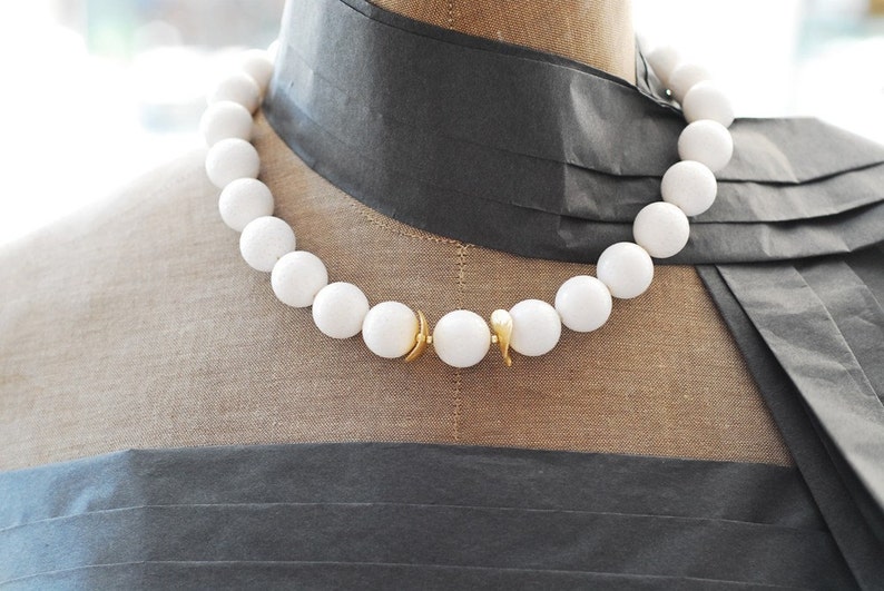 Puede incluir: Un collar de cuentas blancas con un cierre dorado. Las cuentas son grandes y redondas, y el collar est&aacute; en un maniqu&iacute;.