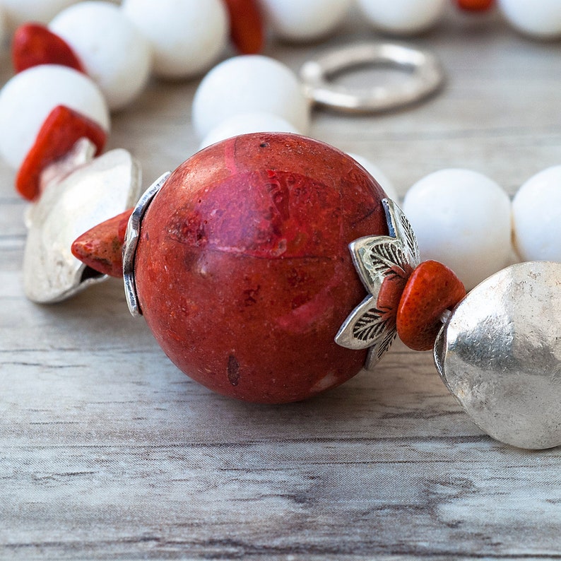 May include: A close-up of a red coral bead necklace with silver accents. The necklace features a large, round red coral bead with a silver leaf design and a smaller red coral bead with a silver charm.