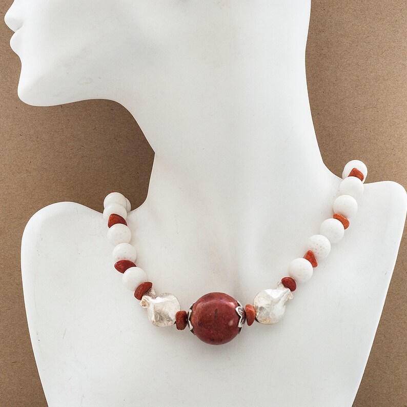 May include: A white and red beaded necklace with a large red stone pendant. The necklace is on a white mannequin.