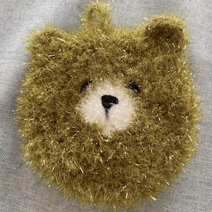 May include: A handmade, round, golden-brown bear-shaped item with a loop for hanging. The bear has a white muzzle, black eyes, and a black nose. The item is made of a fuzzy, textured material with gold metallic threads.