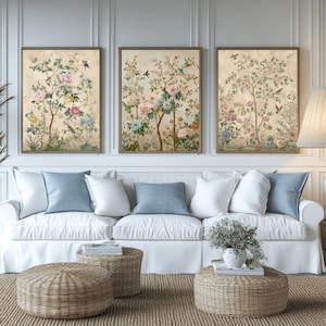 Könnte beinhalten: Drei gerahmte Kunstdrucke mit floralen Motiven im Vintage-Stil hängen über einem weißen Sofa mit blauen und weißen Kissen. Geflochtene Ottomanen und ein Jute-Teppich vervollständigen die Wohnzimmerdekoration. Die Kunstwerke zeigen Vögel und bunte Blumen.