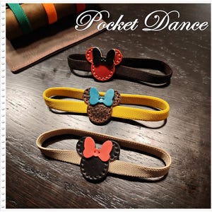 May include: Three elastic bands featuring leather mouse-shaped embellishments with bows. The top band is brown with a red mouse and black bow. The middle is yellow with a brown mouse and blue bow. The bottom is beige with a black mouse and red bow.