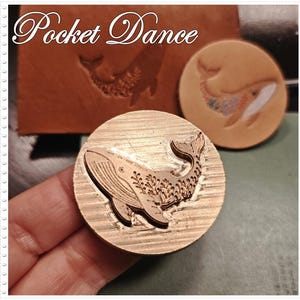 May include: A brass whale stamp with intricate details, held in a hand. The stamp is round and features a whale design with floral accents. The image also shows an embossed whale on leather and a stamped whale.