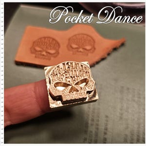 以下が含まれることがあります： 指で持たれた、スカルデザインの金色の金属スタンプ。スタンプは正方形で、「Harley Davidson」の文字がスカルの形にデザインされています。背景には、スタンプされた革片と「Pocket Dance」の文字があります。