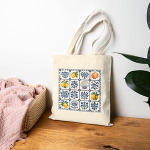 May include: A cream-colored canvas tote bag featuring a square grid pattern of blue and white tiles. The design includes illustrations of yellow lemons, blue floral motifs, seashells, and a small blue fish within the individual tile squares.