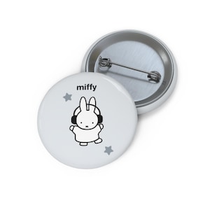 May include: A round white pin-back button featuring a black line drawing of Miffy wearing headphones, with the text miffy printed above the character and two small grey stars positioned on the left and right sides of the design.