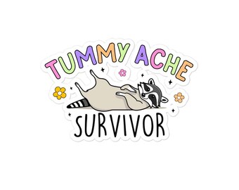 May include: A sticker featuring a cartoon raccoon lying on its back. The text "TUMMY ACHE" is above the raccoon in colorful bubble letters, and "SURVIVOR" is below. Small flowers and stars decorate the design.
