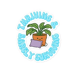 Puede incluir: Una pegatina con un contorno azul y blanco que presenta una planta de dibujos animados en una maceta con una almohada. El texto dice "Thriving & Barely Surviving".