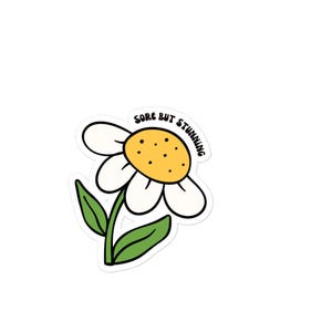 May include: A white daisy sticker with a yellow center and green stem and leaves. The sticker has the words "Sore but Stunning" in a black, rounded font.