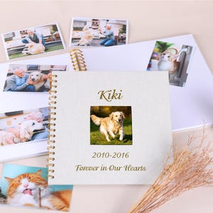 May include: A white fabric photo album with a golden retriever image and the name "Kiki" printed on the cover. The album includes photos of a dog and a cat, with the text "2010-2016 Forever in Our Hearts".