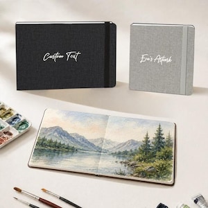 Könnte beinhalten: Ein schwarzes Querformat-Skizzenbuch mit dem Text Custom Text, ein graues quadratisches Skizzenbuch mit dem Text Eva's Artwork und ein offenes Skizzenbuch mit einem Aquarellbild eines Bergsees mit Tannen, begleitet von Pinseln.