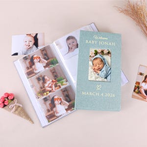 May include: A light blue photo album with the text "Welcome BABY JONAH" and "MARCH 4.2026". The album contains a photo of a baby and several other photos. A small bouquet of red flowers is in the lower left corner.