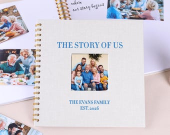 May include: A white linen-textured photo album with gold spiral binding. The cover reads "THE STORY OF US" in blue text, with a family photo in a central window. Below the photo, it says "THE EVANS FAMILY EST. 2026".