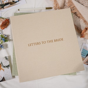 Op de afbeelding: Een beige, met stof bedekt boek met de gouden tekst "LETTERS TO THE BRIDE" op de omslag. Het boek ligt op een groen, getextureerd oppervlak. Een pen en foto's zijn zichtbaar op de achtergrond.