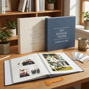 Könnte beinhalten: Zwei Hardcover-Erinnerungsalben mit dem Titel MY SCHOOL YEARS in Creme und Marineblau, wobei eines aufgeschlagen ist und Seiten mit gedruckten Fotos von Schülern in Abschlusskleidung zeigt, neben einem kleinen orangefarbenen Kunststoffwerkzeug.