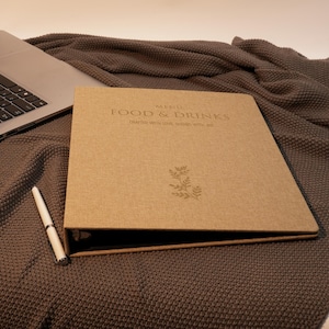 Puede incluir: Un menú marrón con las palabras "FOOD & DRINKS" impresas en la portada, junto con un diseño floral. Una computadora portátil y un bolígrafo están junto al menú sobre una manta marrón texturizada.
